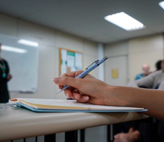 Edital publicado para concurso de ACTs em Santa Catarina - close em mão segurando caneta sobre caderno em sala de aula e professora em pé ao fundo dando aula