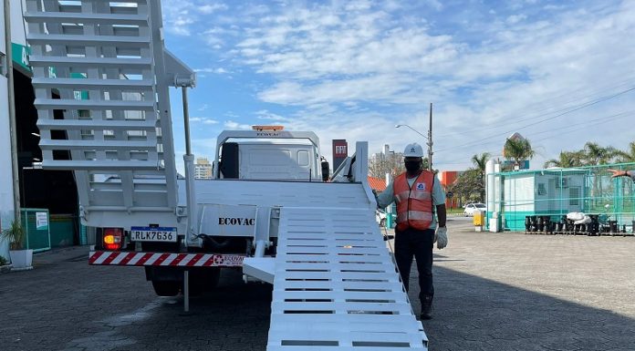 Um caminhão branco aparece de trás, com uma escada estendida e outra recolhida, há um funcionário de colete laranja ao lado. A foto ilustra a notícia de que a casan entregou um lote de equipamentos pesados em quatro superintendências.