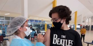 Vacinação contra Covid em adolescentes - rapaz usando camiseta dos beatles olha para enfermeira aplicando dose em seu braço