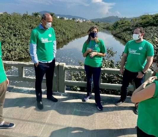 Seis pessoas de camisa verde e usando máscaras conversam em circulo, atrás deles o Rio Capivari. A foto ilustra a ação do programa Trato pelo Capivari, da Casan nos Ingleses, que realizou ação de conscientização com moradores do bairro.