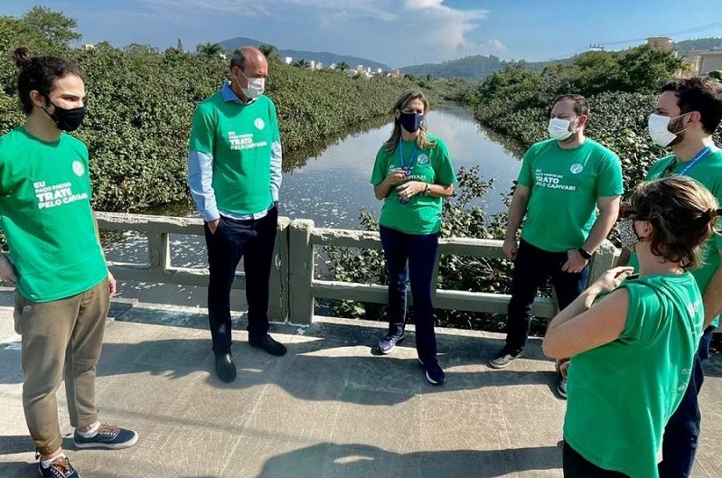 Seis pessoas de camisa verde e usando máscaras conversam em circulo, atrás deles o Rio Capivari. A foto ilustra a ação do programa Trato pelo Capivari, da Casan nos Ingleses, que realizou ação de conscientização com moradores do bairro.