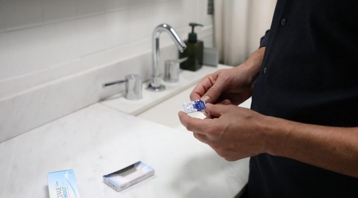 Uma pessoa em frente a uma pia segura nas mãos lentes de contato, com a embalagem apoiada no balcão. A imagem ilustra a notícia de que há uma cartilha com itens para o uso correto das lentes de contato.