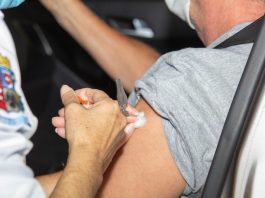 A foto foca na mão de uma pessoa aplicando a vacina em um braço de outra pessoa, sentada em um carro. A imagem ilustra a notícia de que São José vai atualizar os pontos de aplicação da 2ª dose da vacina contra a covid-19