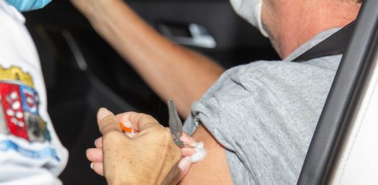 São José atualiza pontos para vacinação da 2ª dose contra a Covid-19 A foto foca na mão de uma pessoa aplicando a vacina em um braço de outra pessoa, sentada em um carro. A imagem ilustra a notícia de que São José vai atualizar os pontos de aplicação da 2ª dose da vacina contra a covid-19