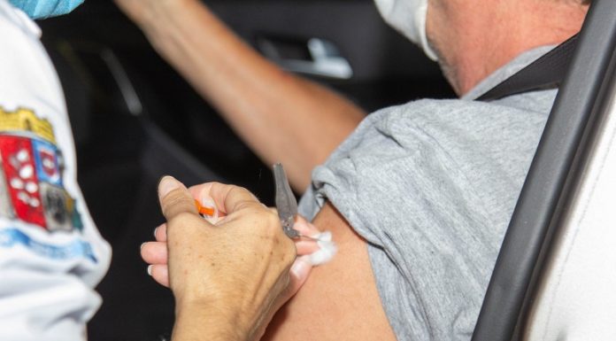 A foto foca na mão de uma pessoa aplicando a vacina em um braço de outra pessoa, sentada em um carro. A imagem ilustra a notícia de que São José vai atualizar os pontos de aplicação da 2ª dose da vacina contra a covid-19