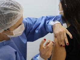 Vacinação de Covid nesta sexta na Grande Florianópolis: locais e horários Profissional de saúde com vestimenta azul, toca e máscara branca, além de unhas pintadas de vermelho, aplica a vacina em um braço, de uma pessoa de cabelos pretos e roupa preta que aparece de costas. A adesão de jovens à vacinação em municípios como Florianópolis, Palhoça e Biguaçu é alta, mas ainda não chega a todos.