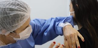 Profissional de saúde com vestimenta azul, toca e máscara branca, além de unhas pintadas de vermelho, aplica a vacina em um braço, de uma pessoa de cabelos pretos e roupa preta que aparece de costas. A adesão de jovens à vacinação em municípios como Florianópolis, Palhoça e Biguaçu é alta, mas ainda não chega a todos.