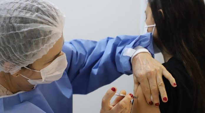 Profissional de saúde com vestimenta azul, toca e máscara branca, além de unhas pintadas de vermelho, aplica a vacina em um braço, de uma pessoa de cabelos pretos e roupa preta que aparece de costas. A adesão de jovens à vacinação em municípios como Florianópolis, Palhoça e Biguaçu é alta, mas ainda não chega a todos.
