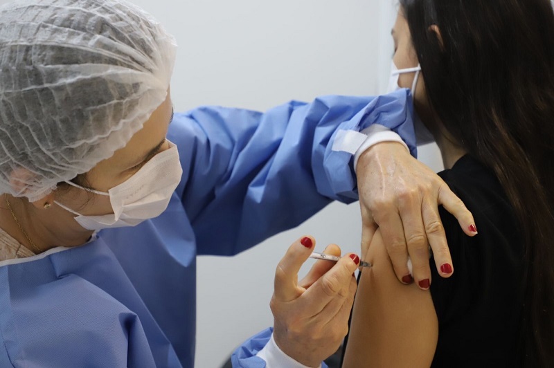 Profissional de saúde com vestimenta azul, toca e máscara branca, além de unhas pintadas de vermelho, aplica a vacina em um braço, de uma pessoa de cabelos pretos e roupa preta que aparece de costas. A adesão de jovens à vacinação em municípios como Florianópolis, Palhoça e Biguaçu é alta, mas ainda não chega a todos.