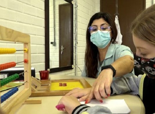 Fundação Catarinense de Educação Especial abre inscrições para concurso público professora de educação especial usando máscara aponta objeto na mesa para aluna usando máscara