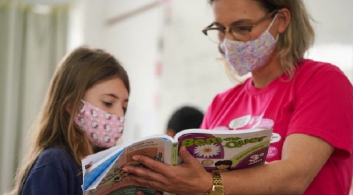 professora e aluna usando mácaras olham para livro didático