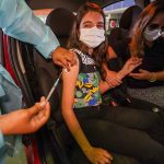 garota bem jovem é vacina contra covid no braço dentro de carro; ela está de máscara e há uma mulher no banco do motorista