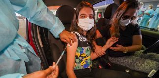 garota bem jovem é vacina contra covid no braço dentro de carro; ela está de máscara e há uma mulher no banco do motorista