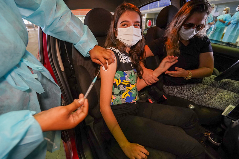 garota bem jovem é vacina contra covid no braço dentro de carro; ela está de máscara e há uma mulher no banco do motorista