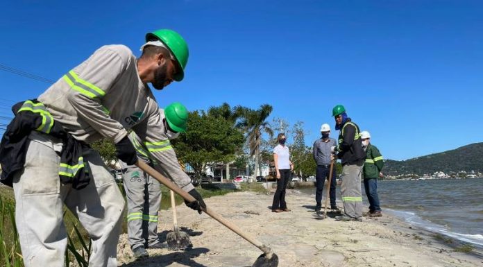 Lagoa da Conceição: Casan inicia recuperação da lagoa de evapoinfiltração - operário usa uma enxada na limpeza da orla da lagoa observado por engenheiros da companhia