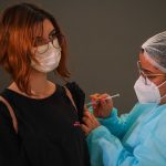 garota usando máscara e óculos recebe injeção no braço dada por enfermeira usando epi - Vacinação em SC: municípios têm disponíveis vacinas suficientes para a primeira dose em 100% dos adolescentes