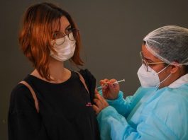 Governo afirma que municípios têm doses suficientes para cobrir adolescentes garota usando máscara e óculos recebe injeção no braço dada por enfermeira usando epi - Vacinação em SC: municípios têm disponíveis vacinas suficientes para a primeira dose em 100% dos adolescentes