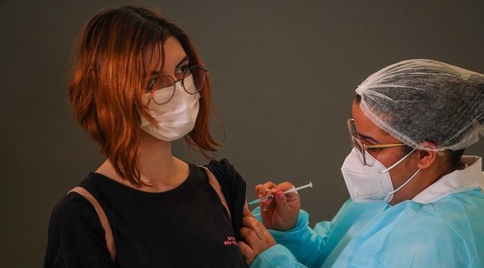 garota usando máscara e óculos recebe injeção no braço dada por enfermeira usando epi - Vacinação em SC: municípios têm disponíveis vacinas suficientes para a primeira dose em 100% dos adolescentes