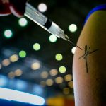 agulha de vacina em frente à tatuagem inscrita fé em braço; vacinação e redução de mortes por covid em sc