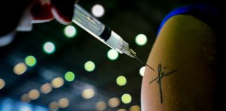 agulha de vacina em frente à tatuagem inscrita fé em braço; vacinação e redução de mortes por covid em sc