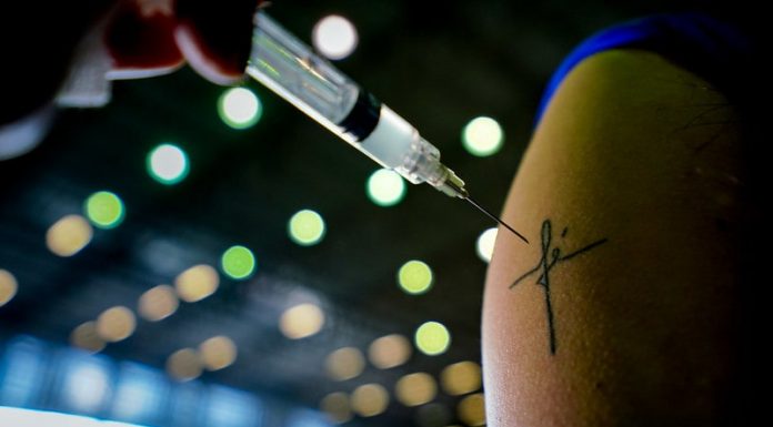 agulha de vacina em frente à tatuagem inscrita fé em braço; vacinação e redução de mortes por covid em sc