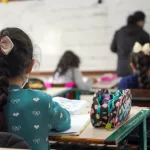 alunas crianças vistas de costas sentadas em carteiras em sala de aula e professora ao fundo