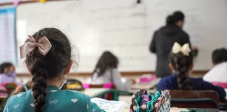 alunas crianças vistas de costas sentadas em carteiras em sala de aula e professora ao fundo