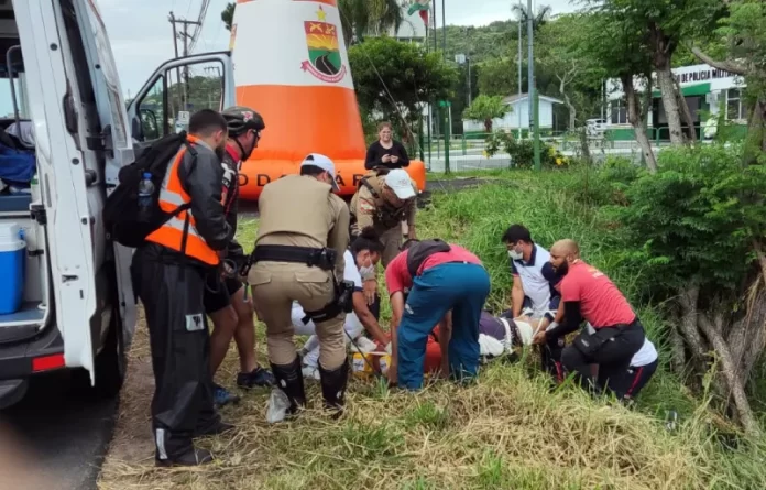 equipes de resgate retirando corpo ao lado de barrando com árvores - Motociclista morre em frente ao posto policial da SC-401