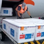 caixas de isopor com o lote de vacinas da janssen sobre carrinho de aeroporto ao lado de compartimento de avião aberto e funcionário manejando as caixas