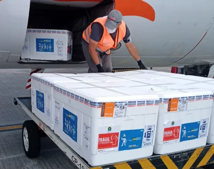 caixas de isopor com o lote de vacinas da janssen sobre carrinho de aeroporto ao lado de compartimento de avião aberto e funcionário manejando as caixas