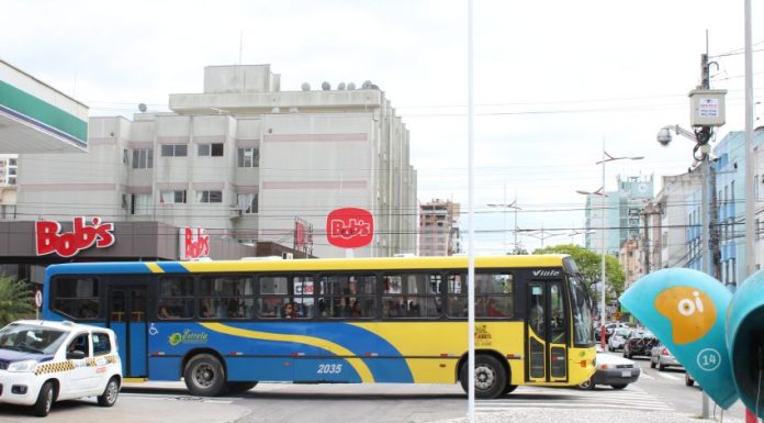 Vista lateral de um ônibus coletivo azul e amarelo cruzando uma avenida com um calçadão no meio