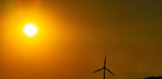 Imagem ilustra calor intenso, com sol forte no horizonte e um aerogerador em silhueta, representando altas temperaturas e clima seco.