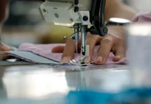 Uma mão feminina está posicionada sobre uma máquina de costura, guiando cuidadosamente um tecido rosa-claro. Os dedos tocam levemente o pano, enquanto a agulha da máquina faz o movimento de costura. A cena transmite concentração e delicadeza, com destaque para os detalhes do tecido e da máquina em funcionamento.