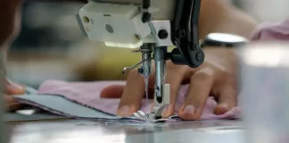 Uma mão feminina está posicionada sobre uma máquina de costura, guiando cuidadosamente um tecido rosa-claro. Os dedos tocam levemente o pano, enquanto a agulha da máquina faz o movimento de costura. A cena transmite concentração e delicadeza, com destaque para os detalhes do tecido e da máquina em funcionamento.