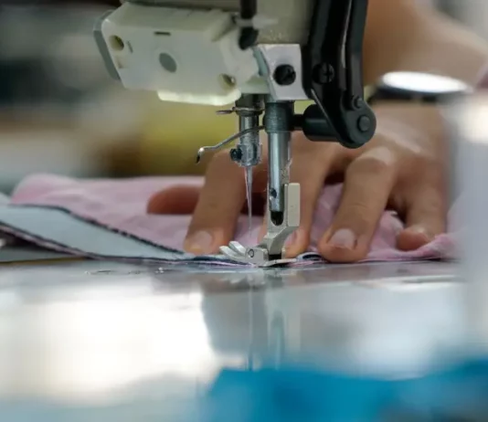 Uma mão feminina está posicionada sobre uma máquina de costura, guiando cuidadosamente um tecido rosa-claro. Os dedos tocam levemente o pano, enquanto a agulha da máquina faz o movimento de costura. A cena transmite concentração e delicadeza, com destaque para os detalhes do tecido e da máquina em funcionamento.