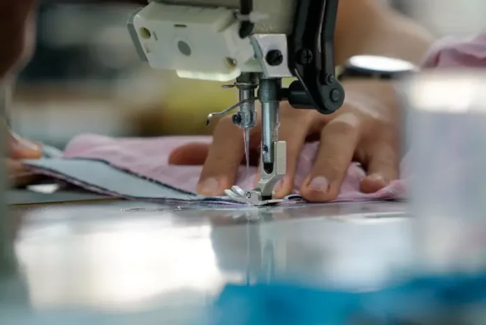 Uma mão feminina está posicionada sobre uma máquina de costura, guiando cuidadosamente um tecido rosa-claro. Os dedos tocam levemente o pano, enquanto a agulha da máquina faz o movimento de costura. A cena transmite concentração e delicadeza, com destaque para os detalhes do tecido e da máquina em funcionamento.