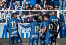 Cinco jogadores do Avaí aparecem em primeiro plano, usando o uniforme oficial azul do time. Estão no gramado da Ressacada, comemorando intensamente a vitória por 2 a 0 sobre o Cuiabá. Alguns se abraçam, outros erguem os braços em sinal de comemoração. Ao fundo, a arquibancada está cheia de torcedores vibrando, muitos em pé, celebrando a conquista com entusiasmo.