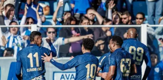 Cinco jogadores do Avaí aparecem em primeiro plano, usando o uniforme oficial azul do time. Estão no gramado da Ressacada, comemorando intensamente a vitória por 2 a 0 sobre o Cuiabá. Alguns se abraçam, outros erguem os braços em sinal de comemoração. Ao fundo, a arquibancada está cheia de torcedores vibrando, muitos em pé, celebrando a conquista com entusiasmo.