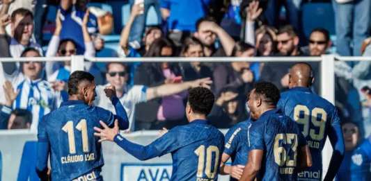 Cinco jogadores do Avaí aparecem em primeiro plano, usando o uniforme oficial azul do time. Estão no gramado da Ressacada, comemorando intensamente a vitória por 2 a 0 sobre o Cuiabá. Alguns se abraçam, outros erguem os braços em sinal de comemoração. Ao fundo, a arquibancada está cheia de torcedores vibrando, muitos em pé, celebrando a conquista com entusiasmo.