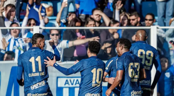 Cinco jogadores do Avaí aparecem em primeiro plano, usando o uniforme oficial azul do time. Estão no gramado da Ressacada, comemorando intensamente a vitória por 2 a 0 sobre o Cuiabá. Alguns se abraçam, outros erguem os braços em sinal de comemoração. Ao fundo, a arquibancada está cheia de torcedores vibrando, muitos em pé, celebrando a conquista com entusiasmo.