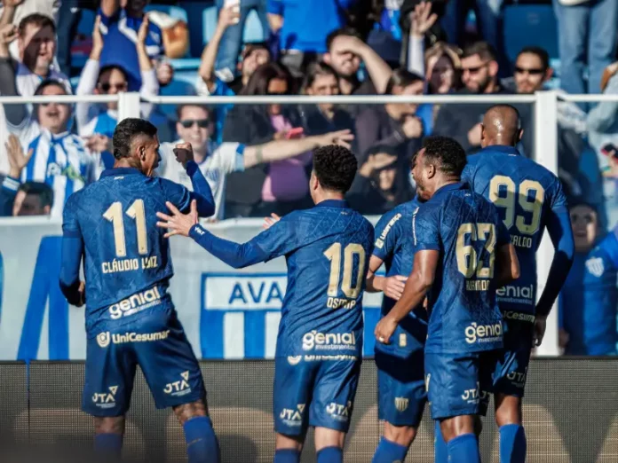 Cinco jogadores do Avaí aparecem em primeiro plano, usando o uniforme oficial azul do time. Estão no gramado da Ressacada, comemorando intensamente a vitória por 2 a 0 sobre o Cuiabá. Alguns se abraçam, outros erguem os braços em sinal de comemoração. Ao fundo, a arquibancada está cheia de torcedores vibrando, muitos em pé, celebrando a conquista com entusiasmo.