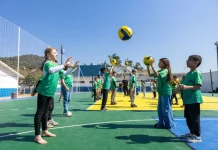 Crianças vestindo camisetas verdes brincam com uma bola na quadra recém-reformada, que tem o chão pintado nas cores verde e amarelo. Elas estão em movimento, sorrindo e se divertindo sob luz natural.