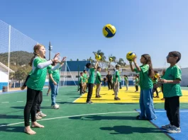 Crianças vestindo camisetas verdes brincam com uma bola na quadra recém-reformada, que tem o chão pintado nas cores verde e amarelo. Elas estão em movimento, sorrindo e se divertindo sob luz natural.