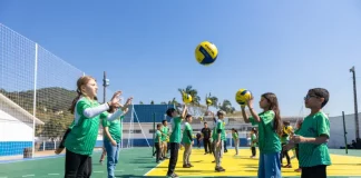 Crianças vestindo camisetas verdes brincam com uma bola na quadra recém-reformada, que tem o chão pintado nas cores verde e amarelo. Elas estão em movimento, sorrindo e se divertindo sob luz natural.