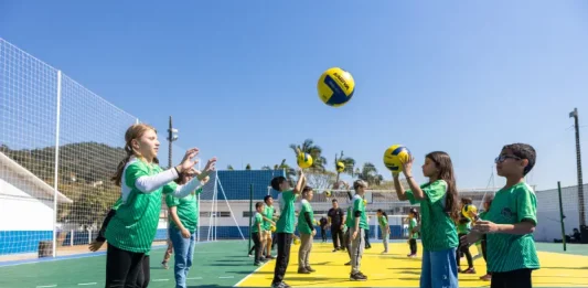 Crianças vestindo camisetas verdes brincam com uma bola na quadra recém-reformada, que tem o chão pintado nas cores verde e amarelo. Elas estão em movimento, sorrindo e se divertindo sob luz natural.