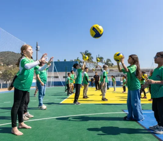 Crianças vestindo camisetas verdes brincam com uma bola na quadra recém-reformada, que tem o chão pintado nas cores verde e amarelo. Elas estão em movimento, sorrindo e se divertindo sob luz natural.