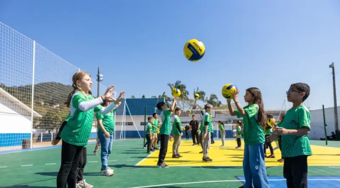 Crianças vestindo camisetas verdes brincam com uma bola na quadra recém-reformada, que tem o chão pintado nas cores verde e amarelo. Elas estão em movimento, sorrindo e se divertindo sob luz natural.