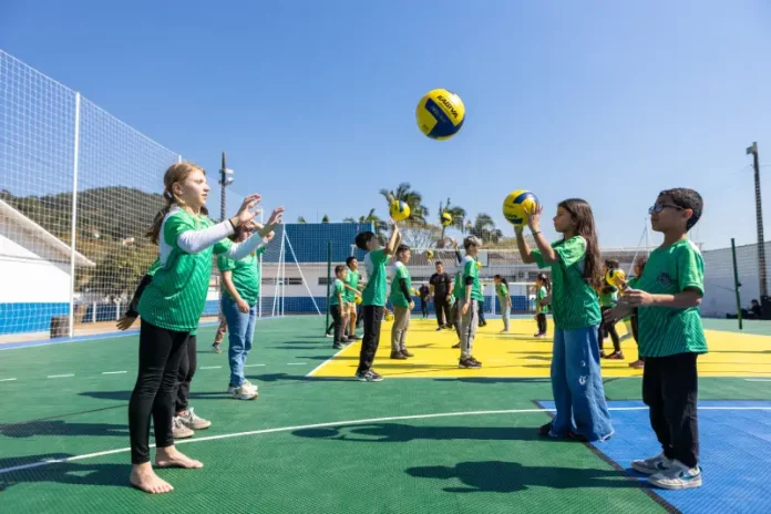 Crianças vestindo camisetas verdes brincam com uma bola na quadra recém-reformada, que tem o chão pintado nas cores verde e amarelo. Elas estão em movimento, sorrindo e se divertindo sob luz natural.