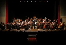 Músicos da Blumenau Filarmônica tocam em um palco com fundo escuro e cortina vermelha ao fundo. Eles estão concentrados em seus instrumentos, enquanto uma plateia de espectadores assiste à apresentação.