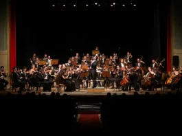 Músicos da Blumenau Filarmônica tocam em um palco com fundo escuro e cortina vermelha ao fundo. Eles estão concentrados em seus instrumentos, enquanto uma plateia de espectadores assiste à apresentação.